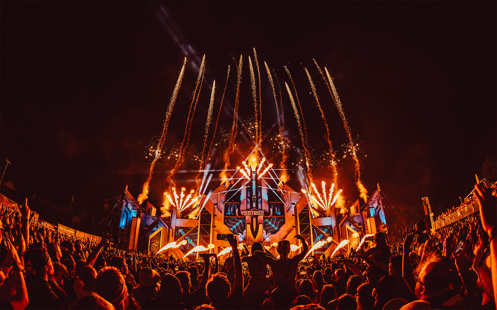 Publikum feiert vor einer Festivalbühne bei Nacht, umgeben von spektakulären Feuerwerkseffekten und Bühnenbeleuchtung beim Electrisize Festival.